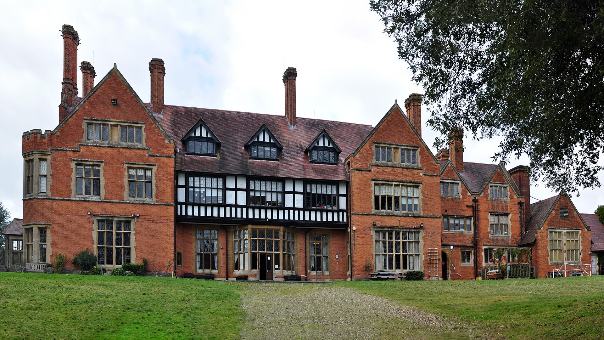 Gentlemans-Country-House_View from the rear of the country manner house from its garden, red brick with stone and lead lined windows, timber framed and white render infill projecting first floor gallery, dormer windows to the roof also timber framed with white render infill, large tall chimney stacks, traditional red clay tiles on steep pitched roofs, with a large tree to the right and sky above