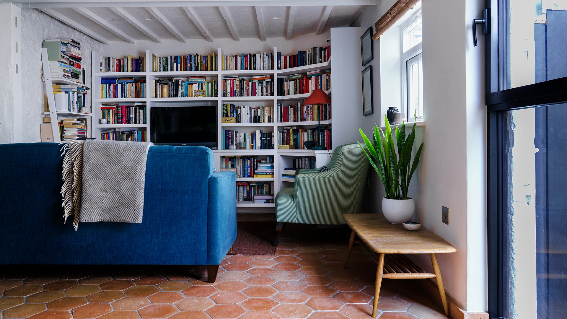 Cosy living room with bookshelves and plants. Interior architecture for a Grade II listed sustainable outbuiling. Living room with clay floor tile finish